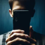 Pessoa esconde o rosto com smartphone preto, segurado por mãos, em fundo azul escuro.